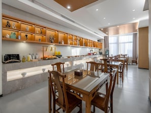 A modern dining area with a long table and chairs, a kitchen with a built-in shelf displaying various items, and a large window providing natural light. - Chonpines Hotel (Yancheng)