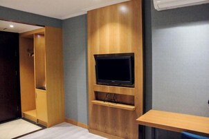 Quarto com televisão integrada, painéis de madeira, uma pequena prateleira e uma escrivaninha.