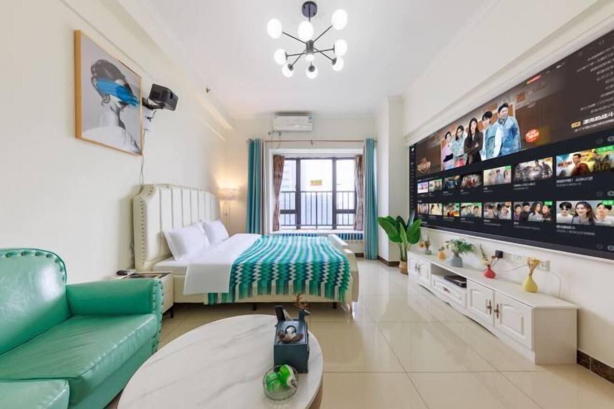 Habitación de hotel con una cama, un sofá verde, una mesa de centro blanca, una pared con televisor y una ventana con cortinas.