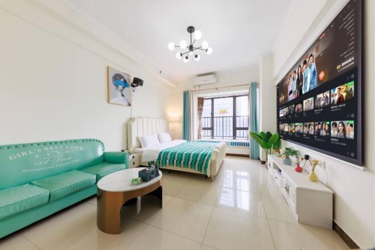 Una habitación de hotel con un sofá verde azulado, una cama con una manta a rayas, una mesa de centro, un televisor montado en la pared y una ventana con cortinas.