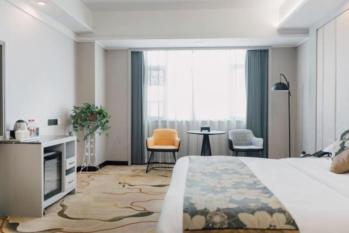 Une chambre d’hôtel avec un lit, un bureau agrémenté d’une plante, deux chaises, une petite table et un lampadaire.