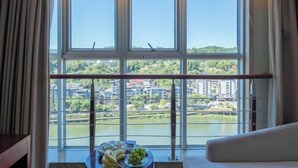 A room with a large window offering a view of a lake and buildings. There is a table with fruits and a sofa.