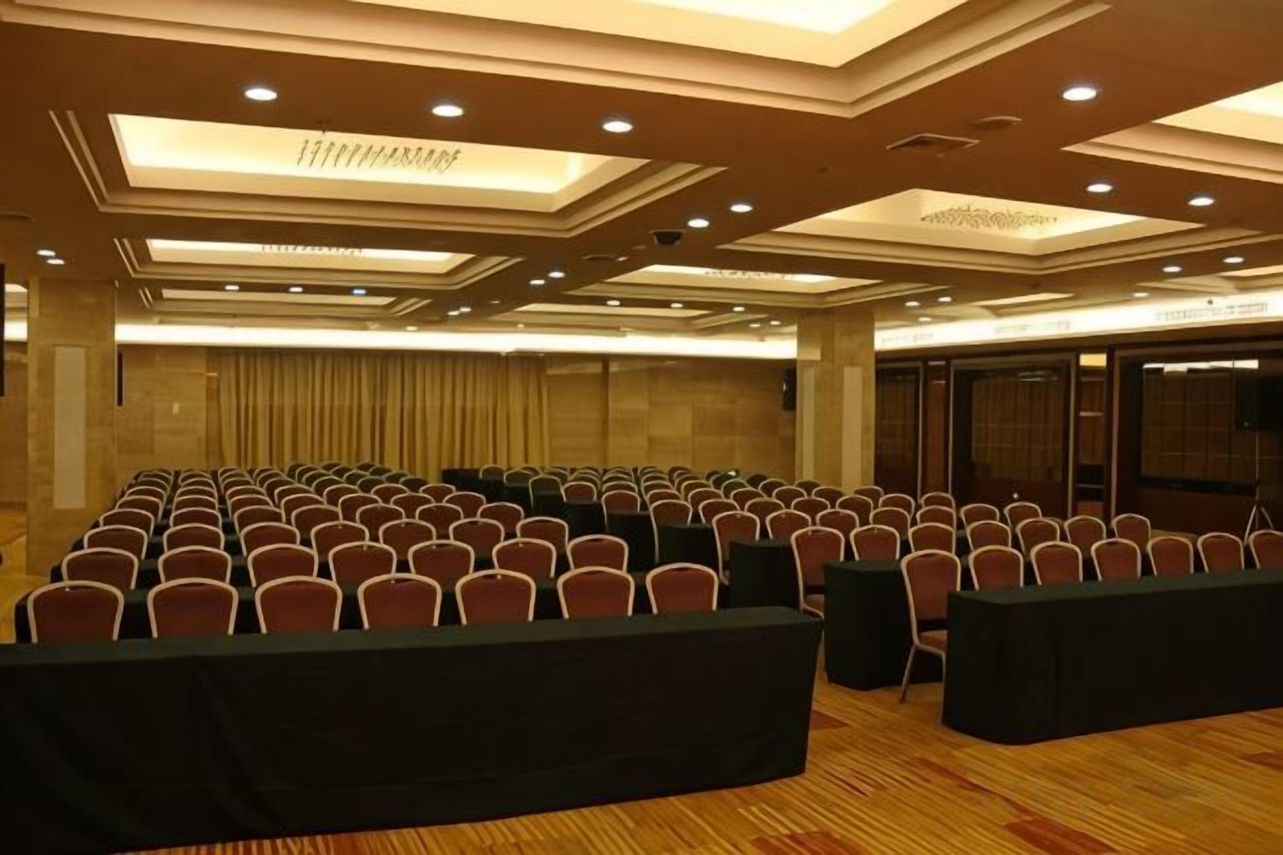 Un salón de conferencias con filas de sillas y mesas, un techo alto con iluminación empotrada y suelo alfombrado.