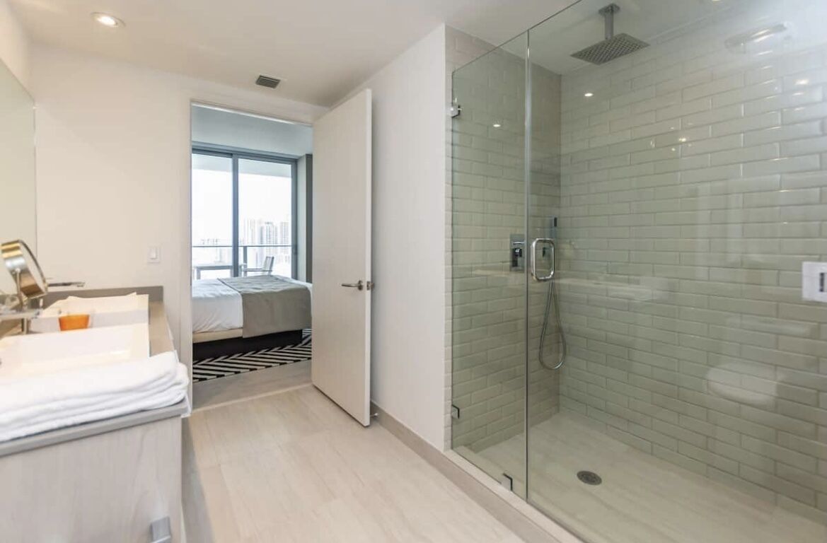 Une salle de bain d’hôtel moderne dotée d’une douche en verre, d’un lit visible dans la pièce adjacente et d’une grande fenêtre laissant entrer la lumière naturelle.