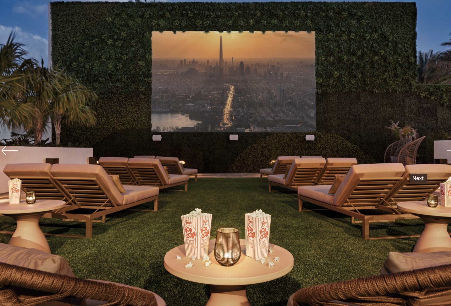 Un salón en la azotea con sillones, una mesa con palomitas y una bebida, y una pantalla grande que muestra vistas de la ciudad.