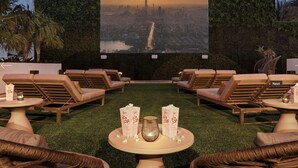 Un salón en la azotea con sillones, una mesa con palomitas y una bebida, y una pantalla grande que muestra vistas de la ciudad.