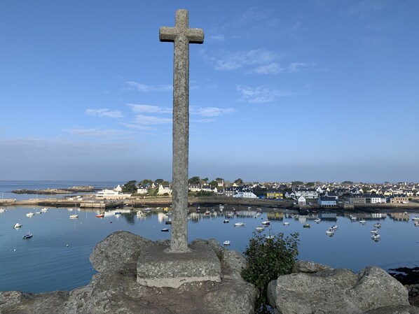 Una cruz de piedra en un pueblo costero con un puerto deportivo y casas al fondo.