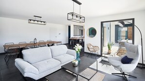 Un salon moderne avec un canapé, une table basse et des fauteuils. On y trouve également un coin repas avec une longue table et des chaises. La pièce est dotée d’une grande fenêtre offrant une vue sur l’extérieur.