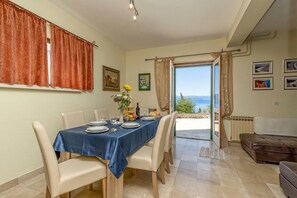 Un'area pranzo con tavolo imbandito per quattro persone, vista sul mare attraverso una porta e un divano sullo sfondo.