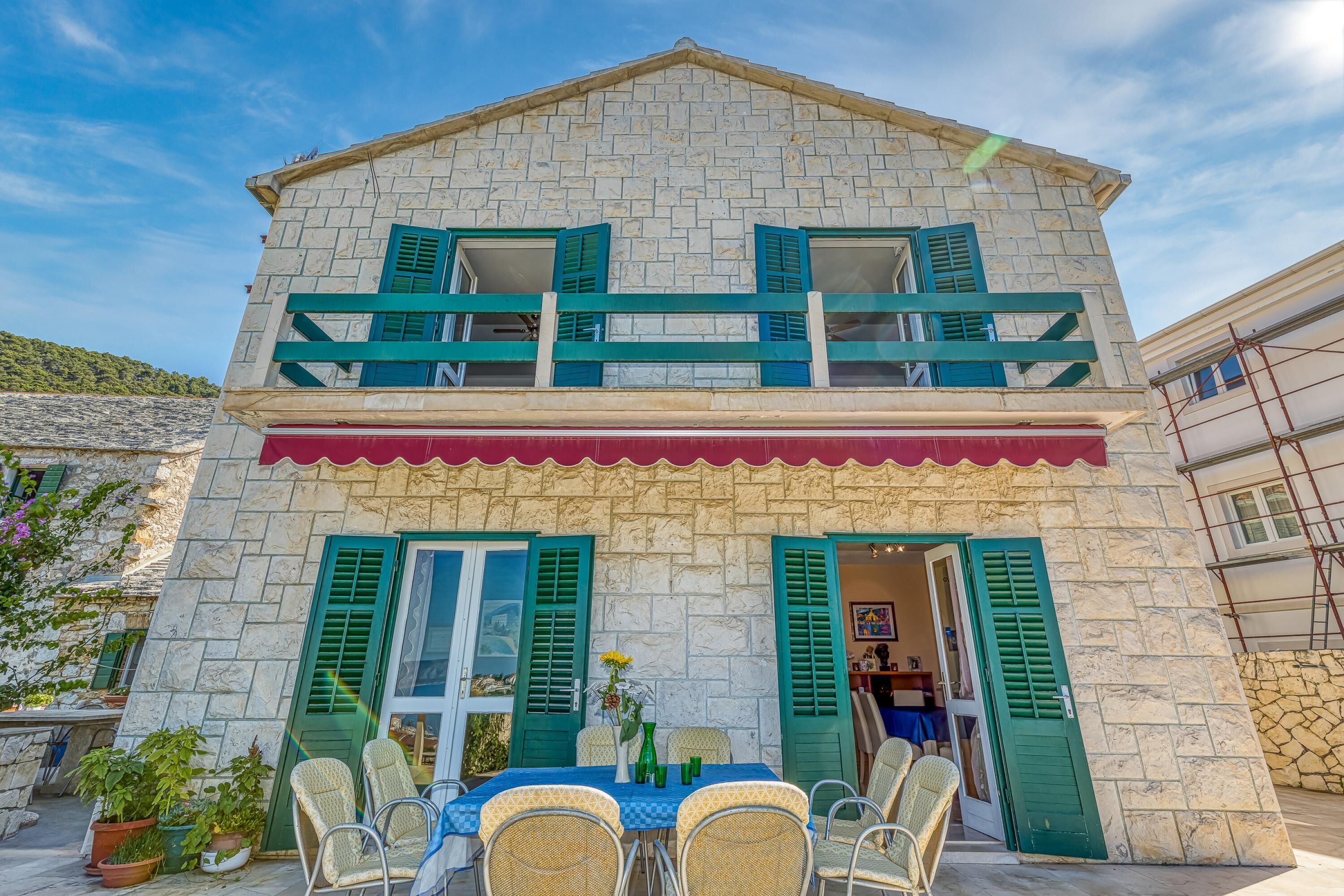 A two-story stone building with green shutters, a balcony, and a dining area with wicker chairs and a table.