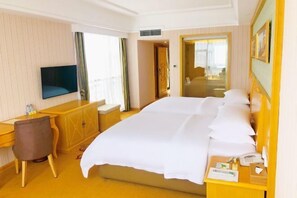 A hotel room with a large bed, a desk with a chair, a television, and a bedside table with a phone and some items. - Biying Grand Hotel (Lingshan)