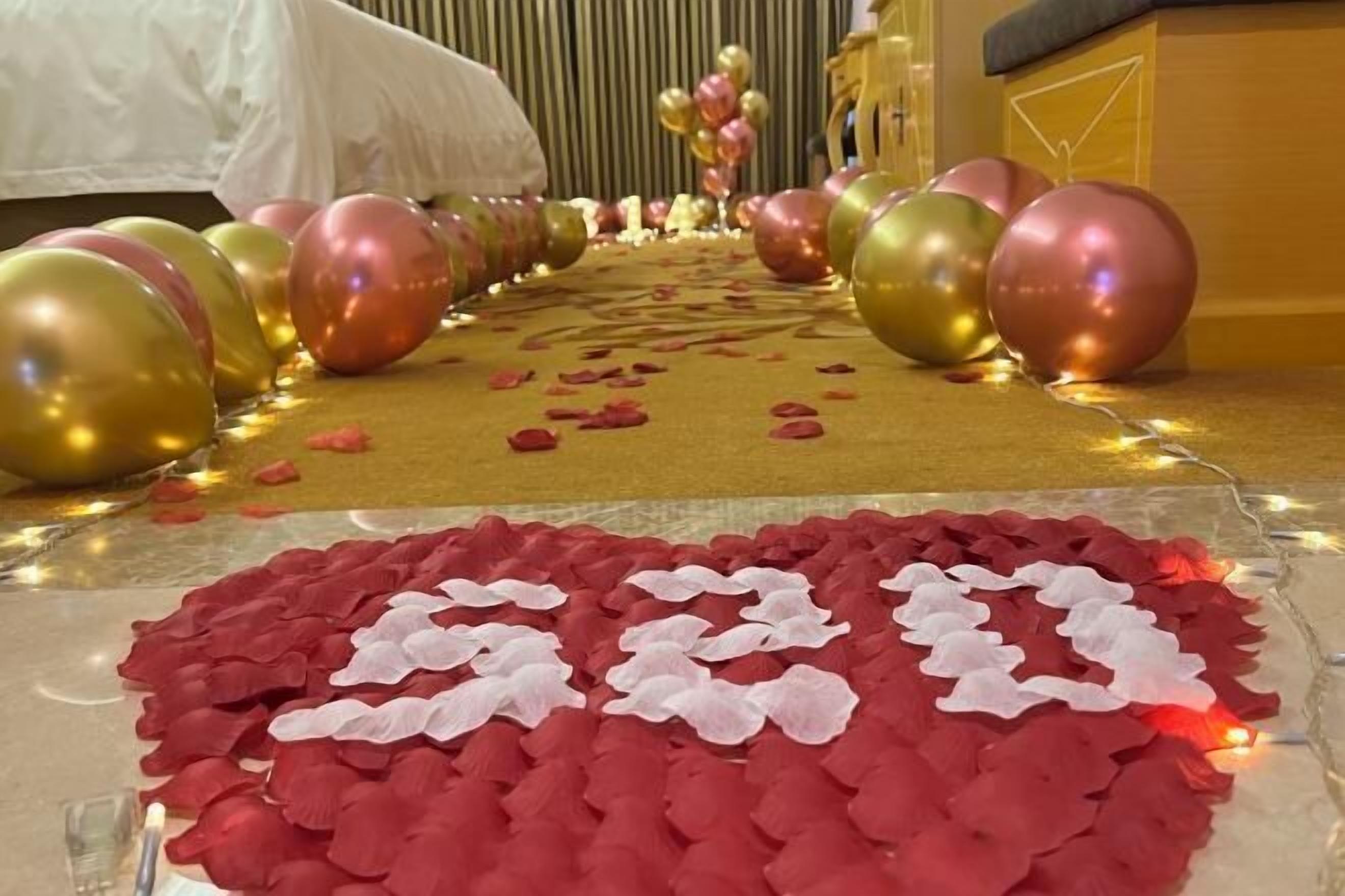 Ein Zimmer mit einem Bett, einer dekorativen Ballon-Anordnung und einem Teppich mit Rosenblättern.