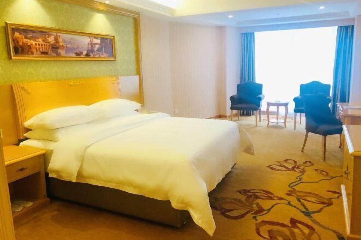 A hotel room with a large bed, a desk, two chairs, and a painting on the wall.