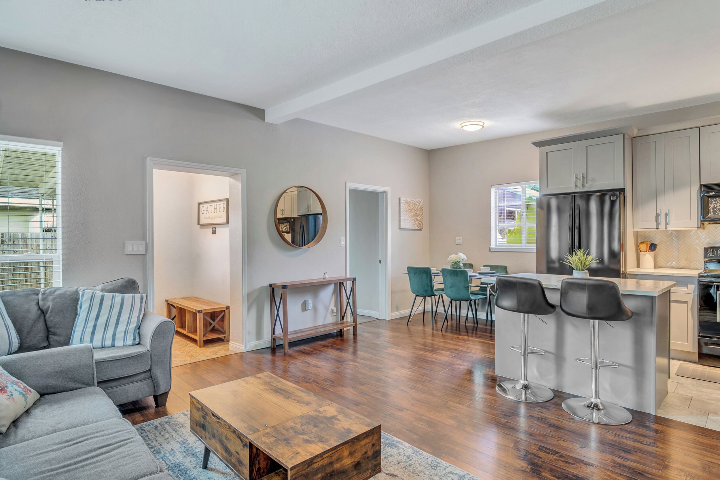 A spacious living room with a grey sofa, wooden coffee table, and a dining area with teal chairs.