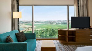 A hotel room with a large window offering a view of a green landscape and cityscape.