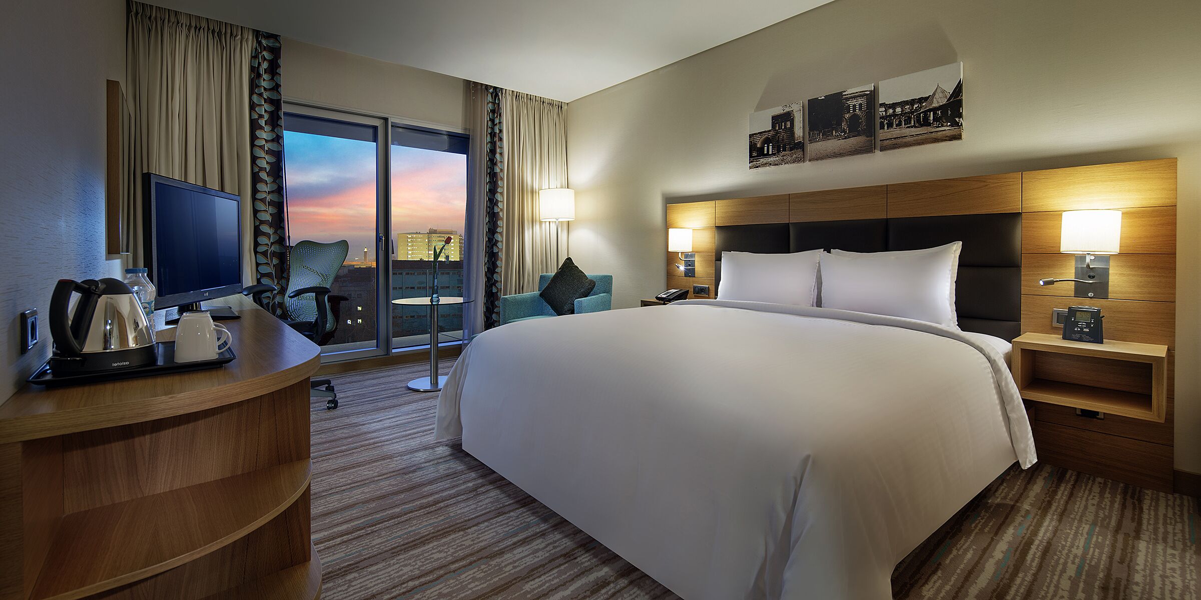 A hotel room with a large bed, a desk with a kettle, a chair, a TV, and a view of the cityscape.