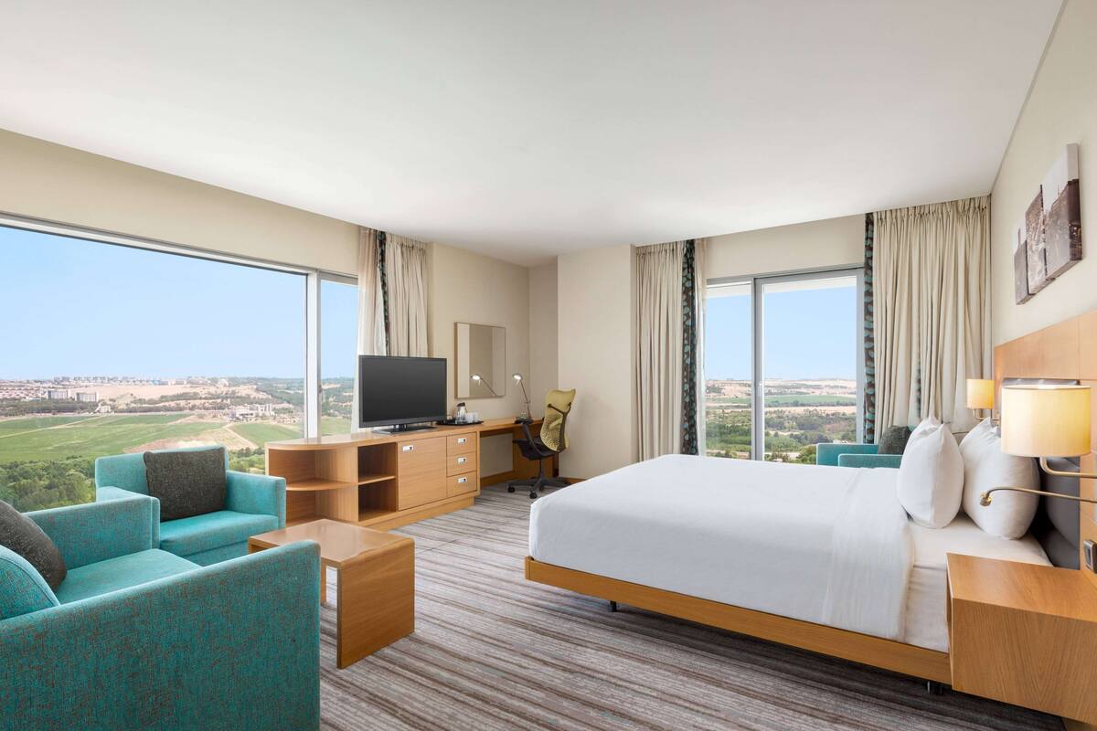 Une chambre d’hôtel avec un grand lit, un bureau avec un ordinateur, un canapé et une vue sur un paysage verdoyant.