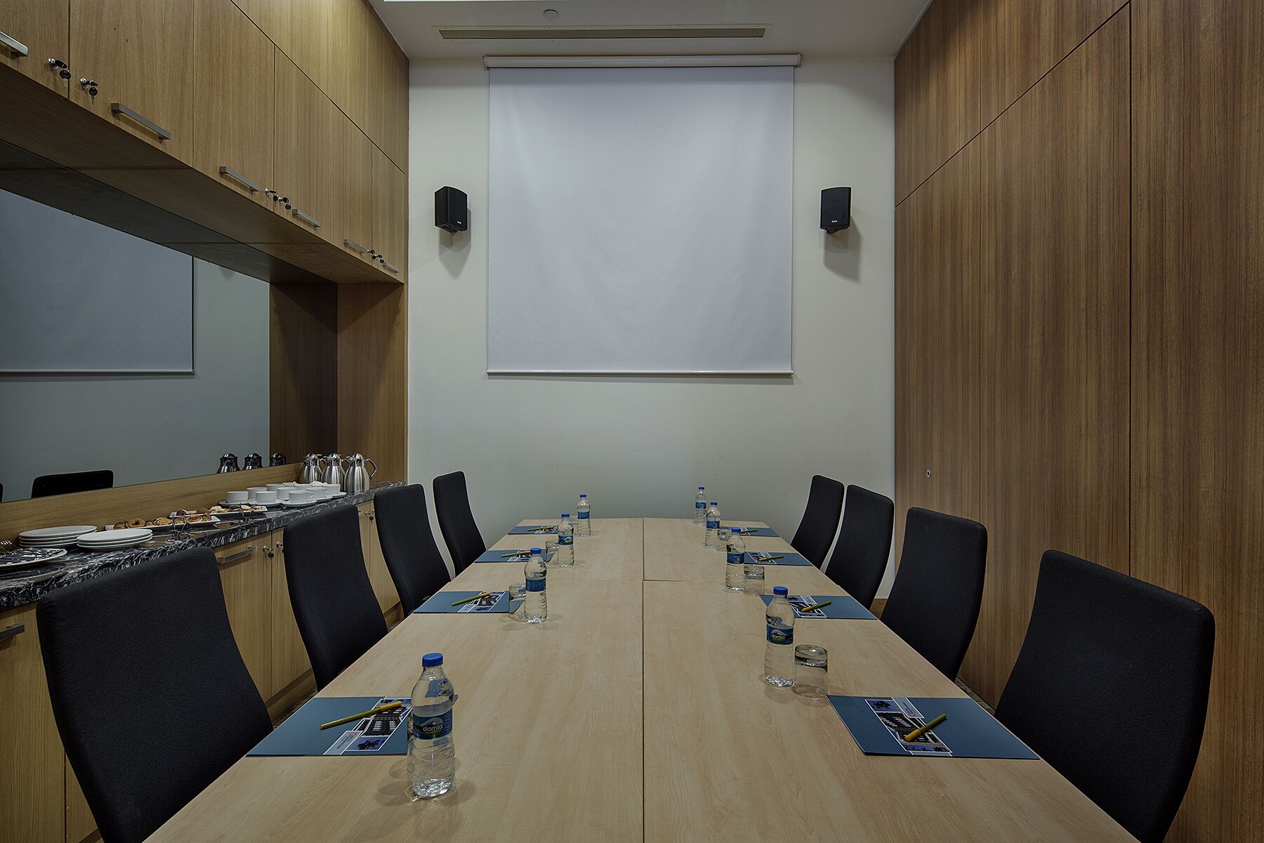Une salle de conférence avec une longue table, des chaises noires, des bouteilles d’eau et un écran de projection.