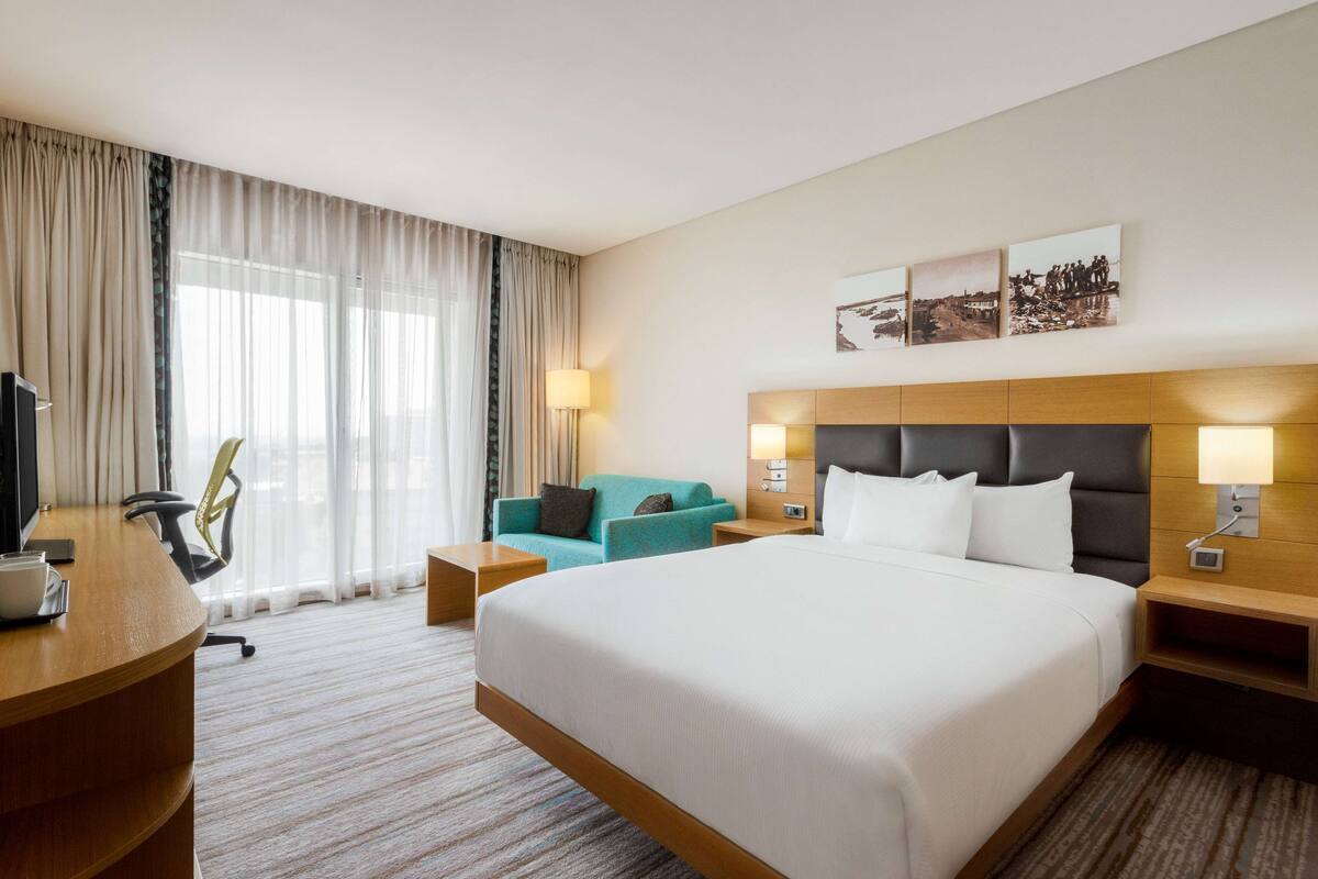 Une chambre d’hôtel avec un grand lit, un bureau avec une chaise, un canapé et une télévision. Il y a des lampes fixées au mur et une fenêtre avec des rideaux.
