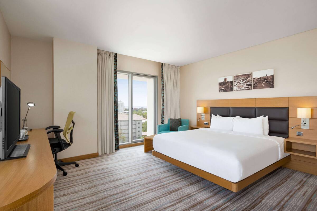 Une chambre d’hôtel avec un grand lit, un bureau avec un ordinateur, une chaise et une vue sur la ville par la fenêtre.