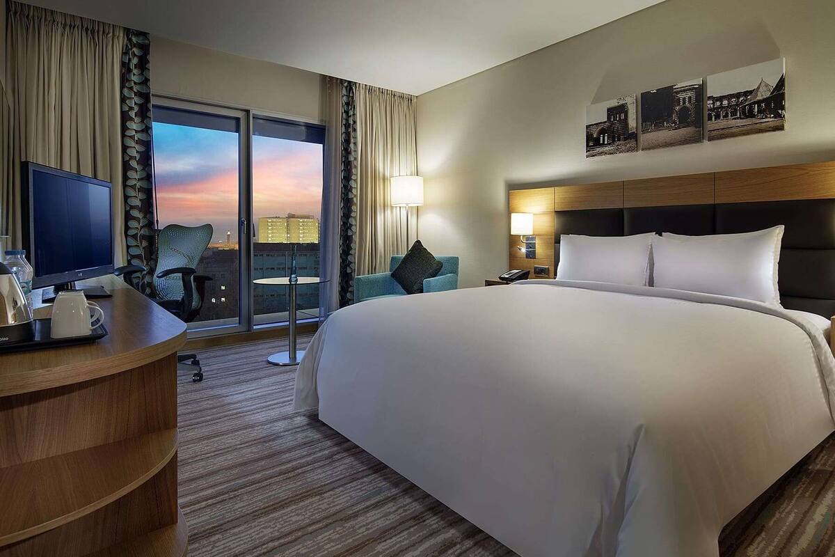 Une chambre d’hôtel avec un grand lit, un bureau avec un ordinateur, une chaise, une télévision et une vue sur le paysage urbain par la fenêtre.