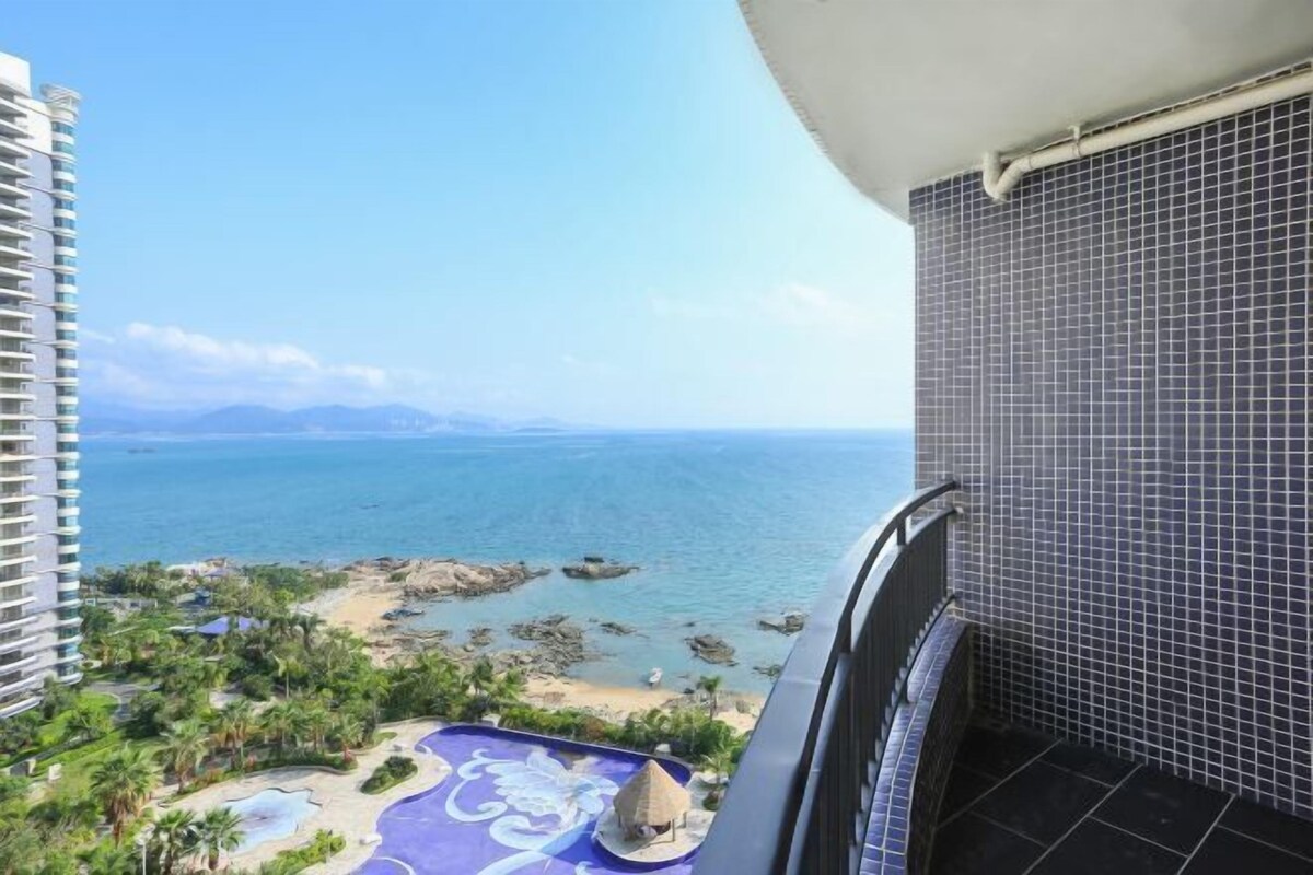 A balcony with a view of a beach and ocean, a tiled wall, and a tropical garden.