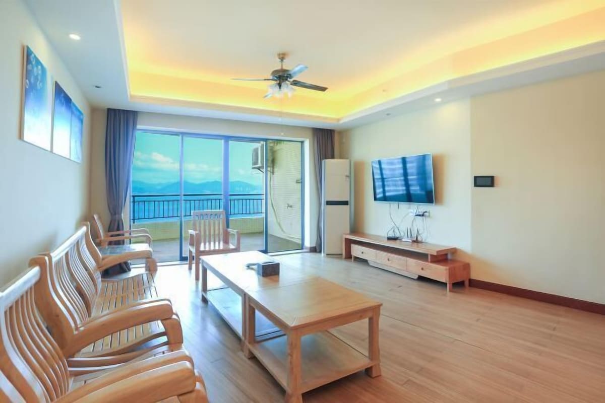 Um quarto espaçoso com piso de madeira, ventilador de teto, uma televisão grande e varanda com vista.