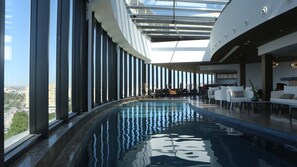 A modern indoor swimming pool with a city view, large windows, and contemporary furniture. - ASBU Hotel (Tunis)