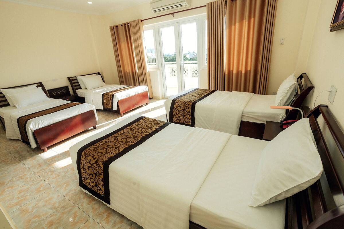 Een hotelkamer met vier bedden op een rij, een balkon met gordijnen en uitzicht op de omgeving.