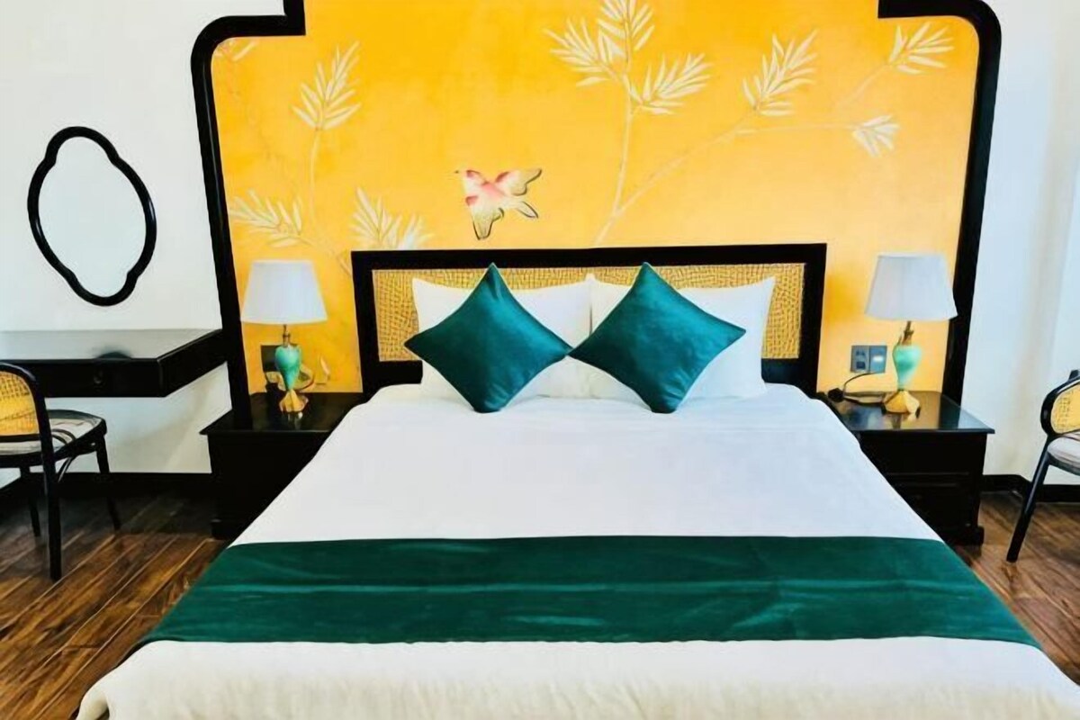 Ein Bett mit weißer und grüner Bettdecke, zwei türkisfarbenen Kissen und einer gelben Wand mit Blättermotiven.