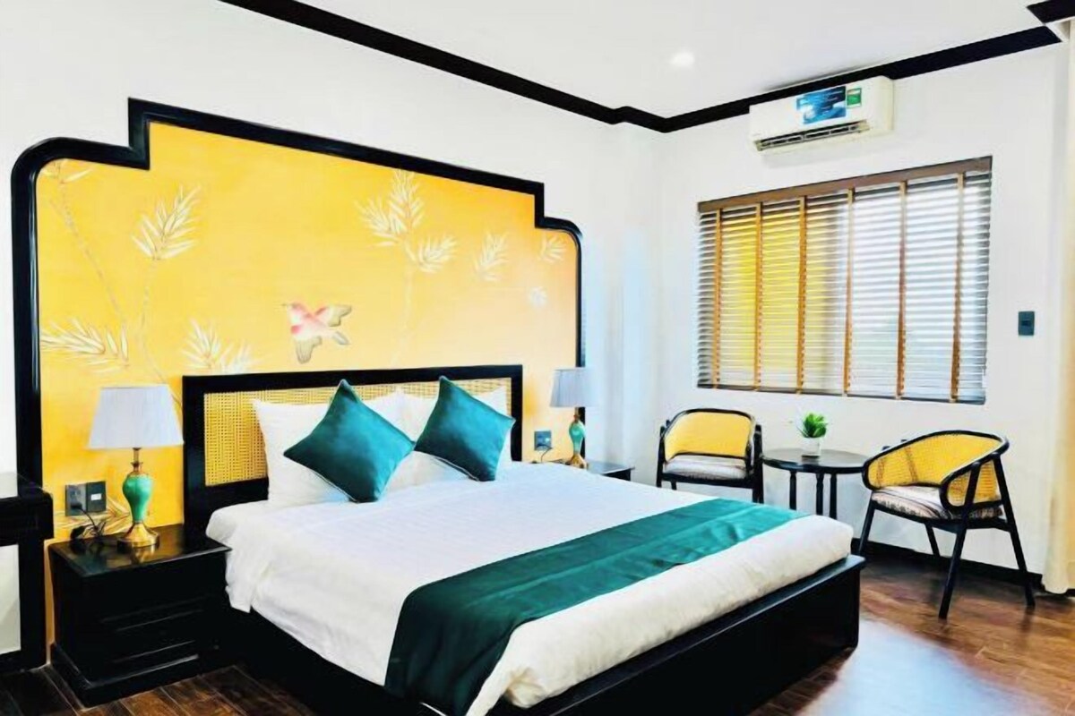Une chambre d’hôtel avec un grand lit, deux chaises, une petite table et une fenêtre avec des stores.