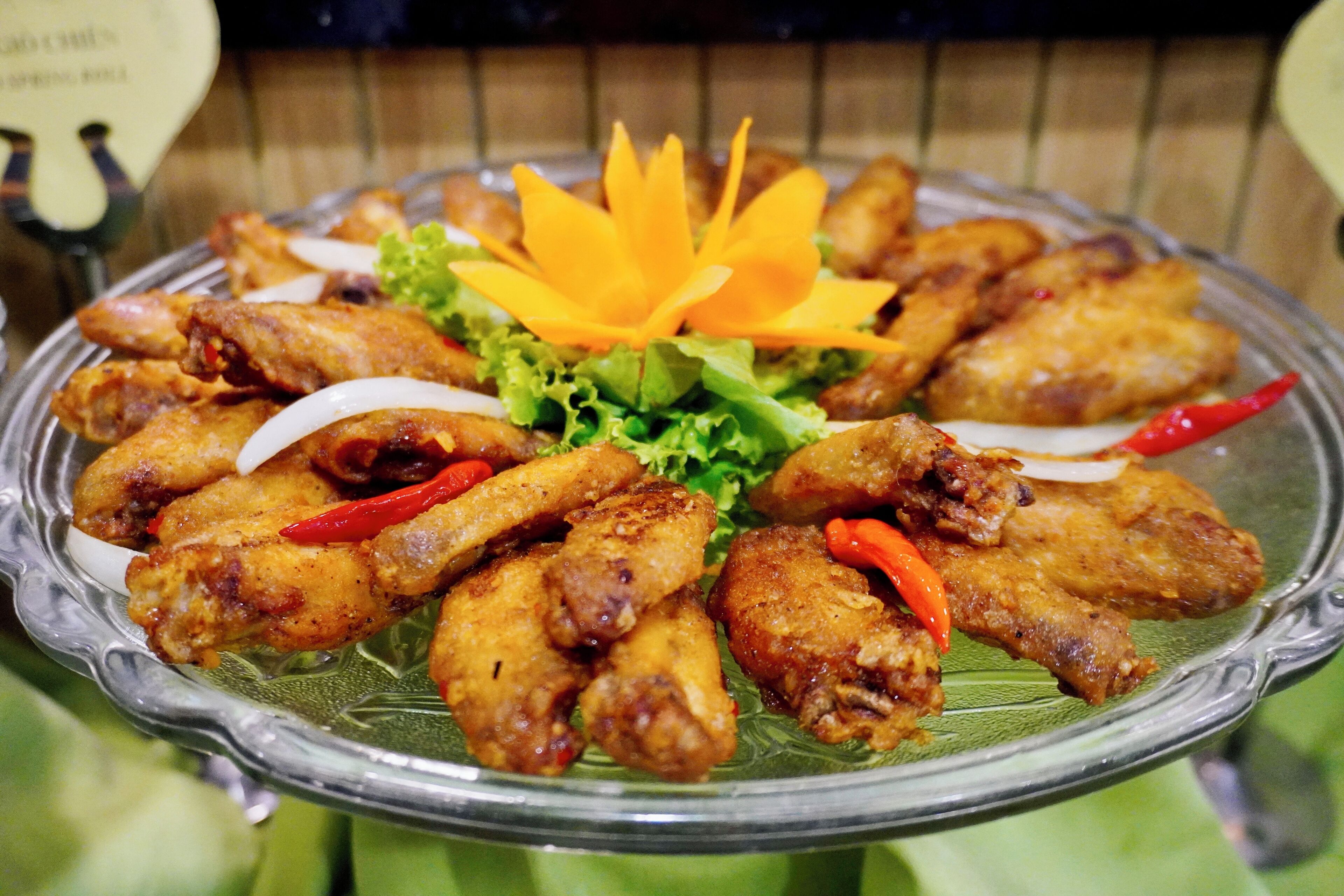 Un plato de vidrio con alitas de pollo fritas, adornadas con lechuga, chiles rojos y una flor amarilla decorativa.