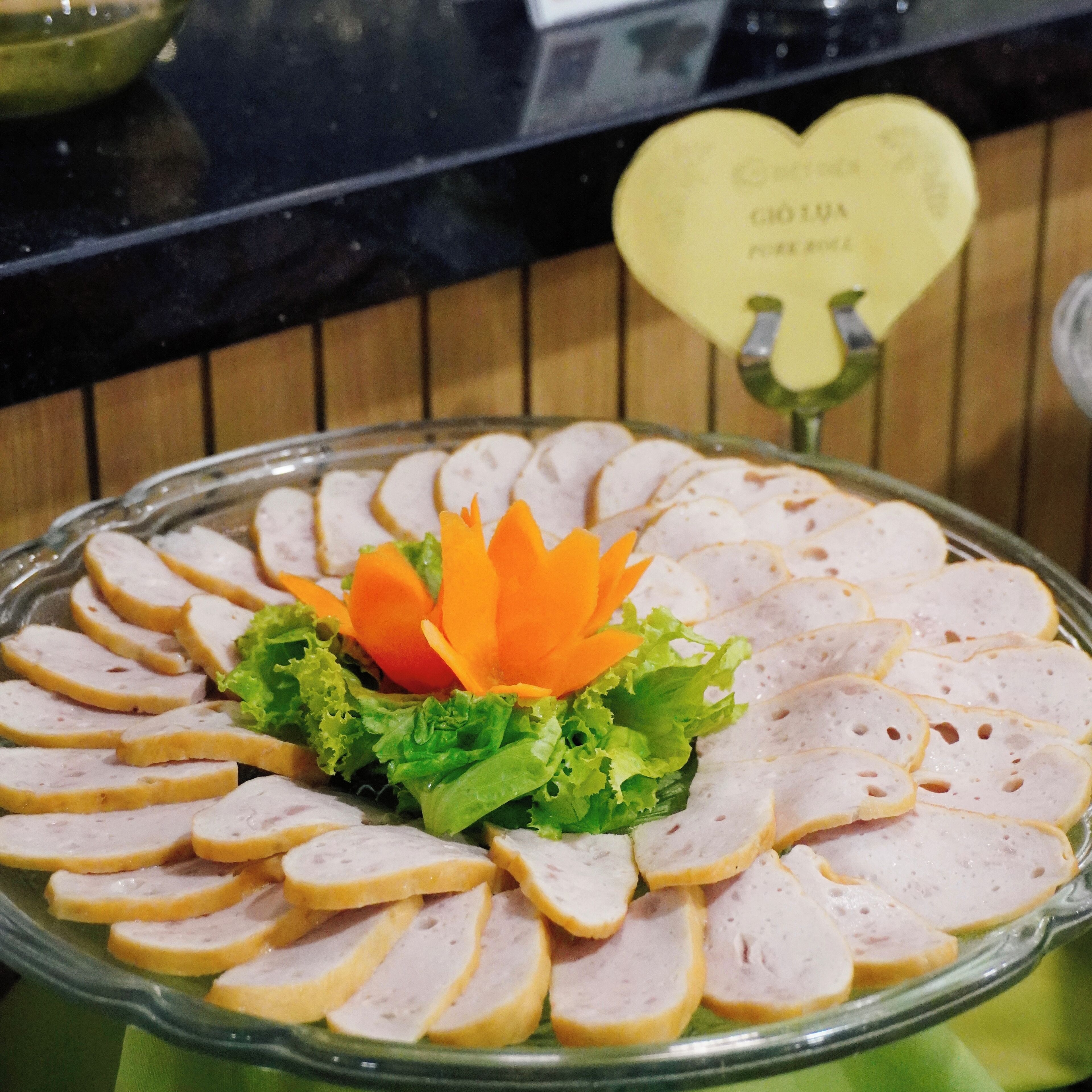 A glass platter with sliced meat arranged in a circular pattern, garnished with lettuce and orange carrot flowers, set on a green cloth.