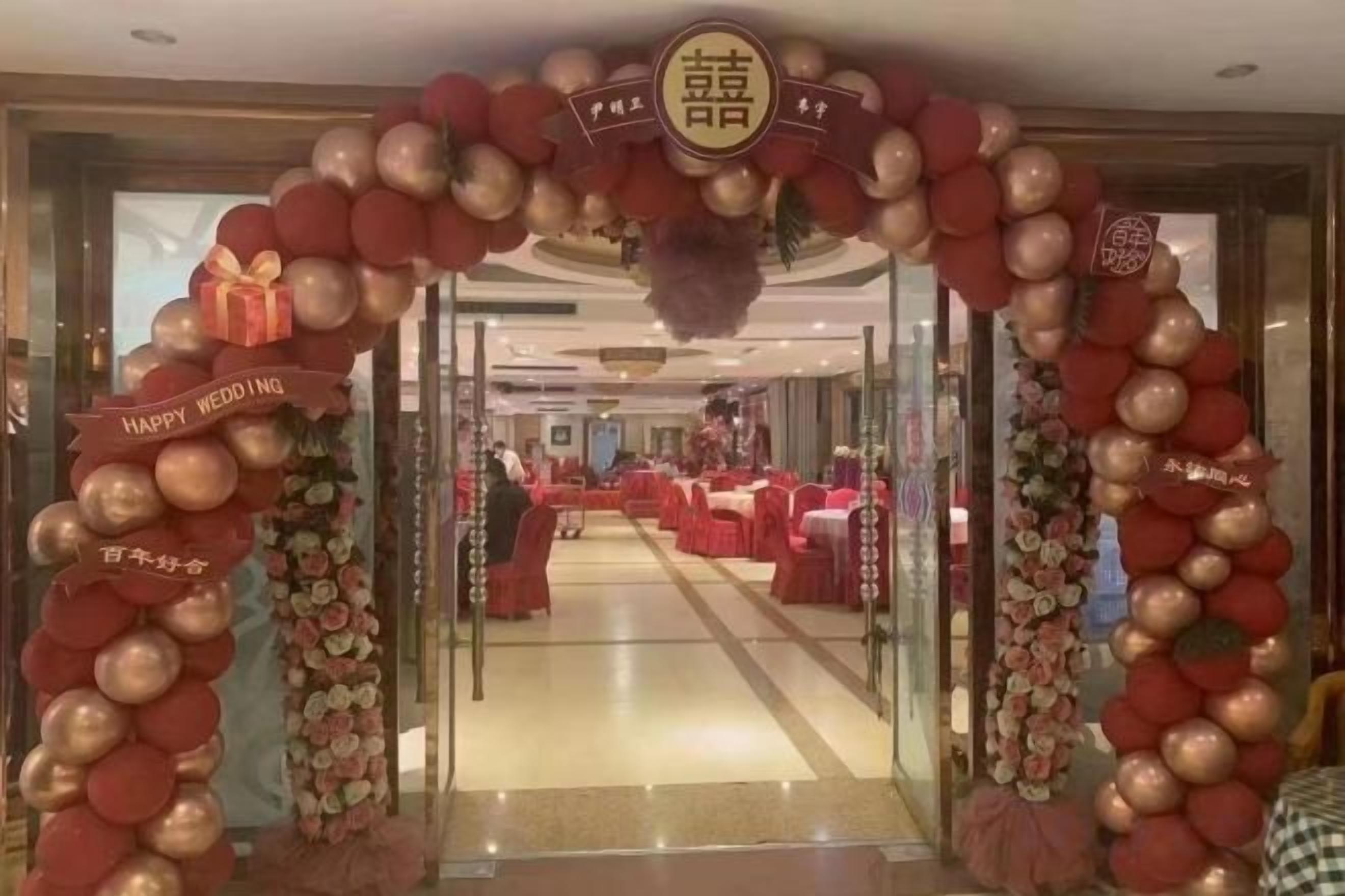 A wedding reception area with a red and gold balloon arch, tables set for guests, and a banner that reads 'HAPPY WEDDING'.