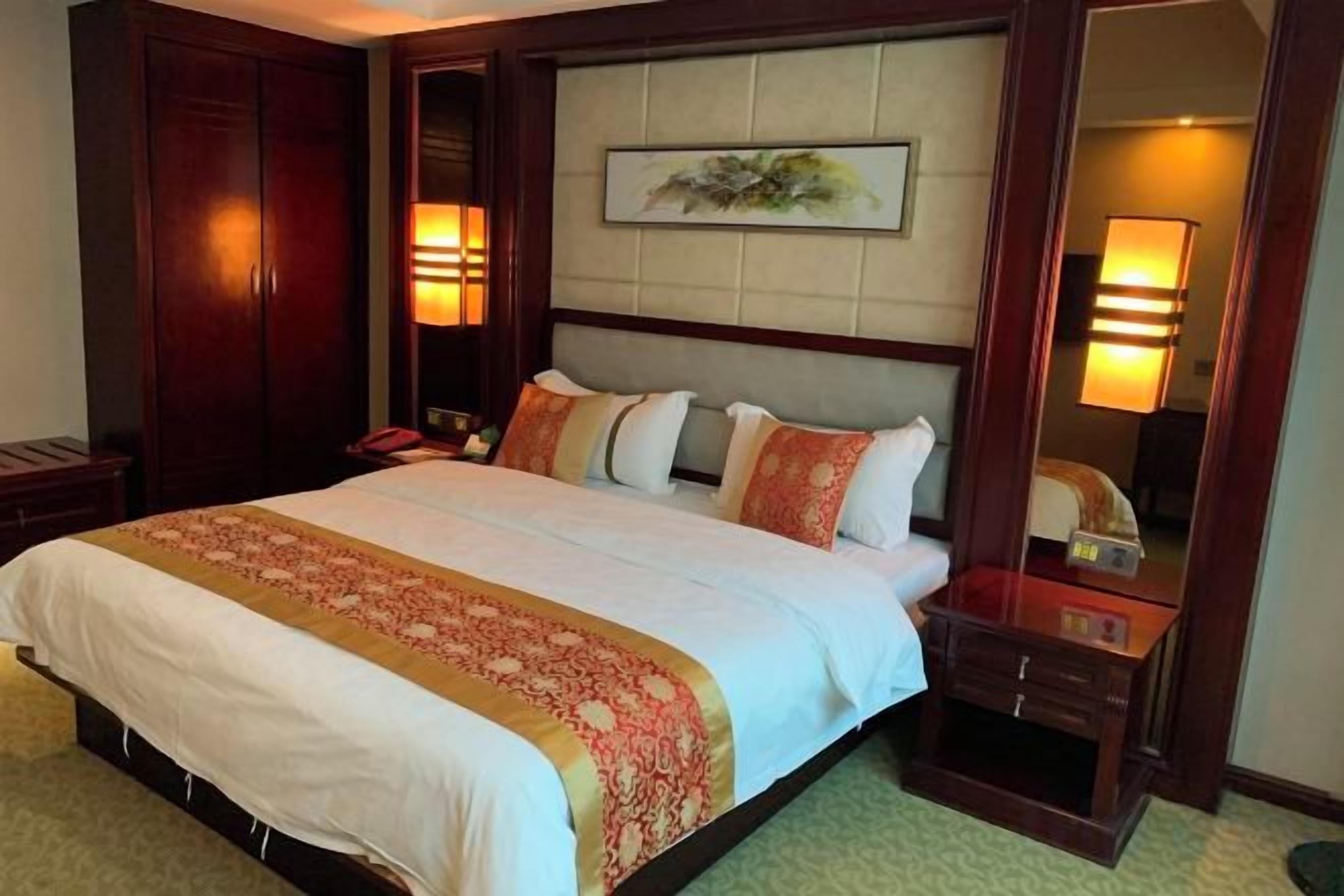 A hotel room with a large bed, a headboard with a mirror, bedside lamps, and a wooden wardrobe.