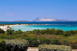 A coastal landscape with a beach, clear blue sea, and a mountain in the background.