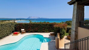Uma piscina com vista para o mar e montanhas, um pilar de pedra e uma cadeira.