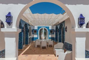 Un espace extérieur aménagé avec une zone salon couverte, comprenant une table ronde et des chaises, entouré de murs blancs ornés de mosaïques bleues, et agrémenté de deux lanternes bleues fixées aux murs.