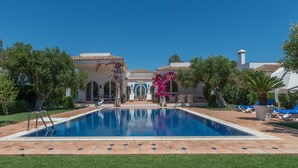 Une piscine dotée d’une terrasse carrelée, entourée de verdure et bordée d’un bâtiment orné d’arches et d’une cheminée.