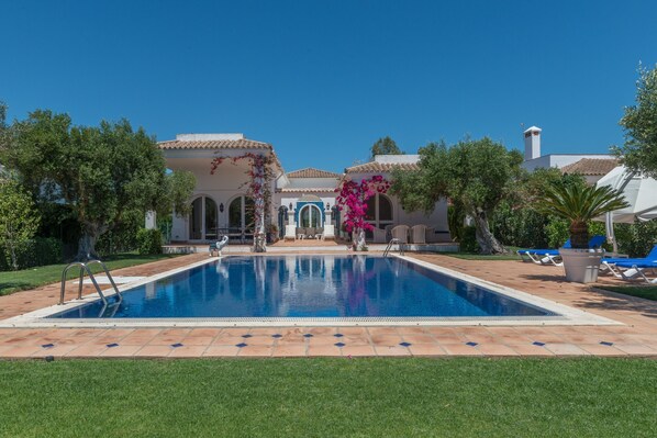 Une piscine dotée d’une terrasse carrelée, entourée de verdure et bordée d’un bâtiment orné d’arches et d’une cheminée.