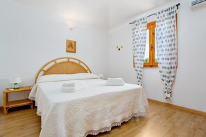A bedroom with a wooden bed, white linens, a bedside table with a lamp, and a window with patterned curtains.