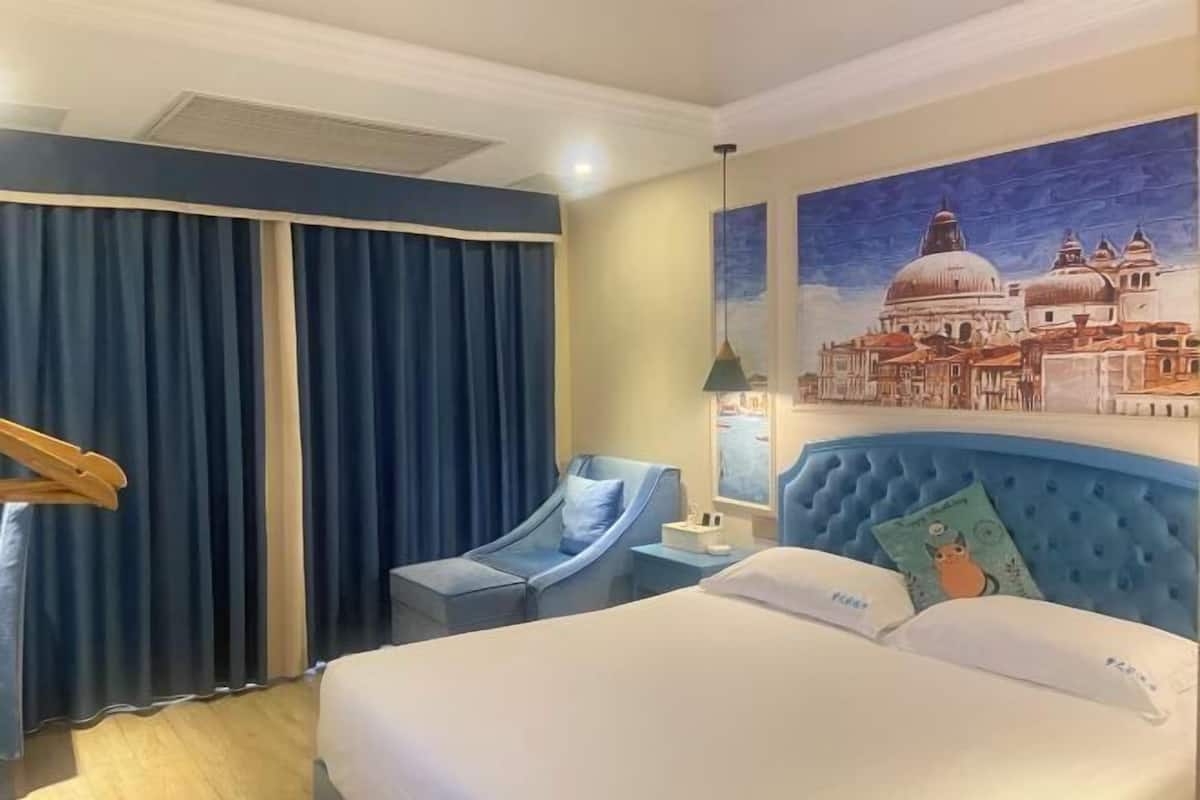 Ein Hotelzimmer mit einem Bett, einer blauen Kopfteil, einem Schreibtisch mit einer Lampe, einem Sessel und einem Stadtbild an der Wand.