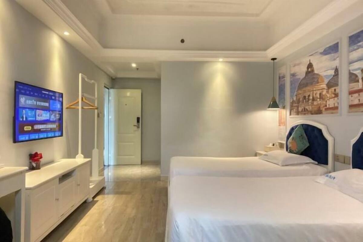 Ein Hotelzimmer mit zwei Betten, einem an der Wand befestigten Fernseher und einem Flur, der in ein weiteres Zimmer führt.