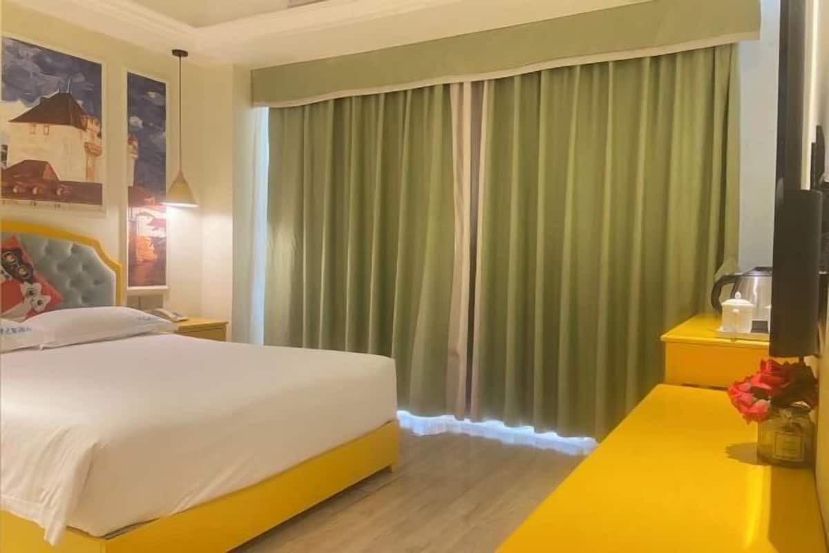 Ein Hotelzimmer mit einem Bett, einem Nachttisch, einem Tisch mit Blumenvase und einem großen Fenster mit Vorhängen.