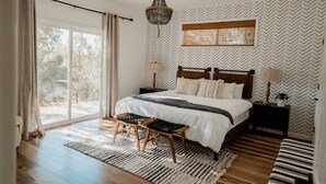 A bedroom with a large window, a bed with a wooden headboard, two bedside tables with lamps, a rug, and a hanging light fixture. - Sunset Glow *Hidden Meadow*BBQ HotTub by Casa Oso (Oakhurst)