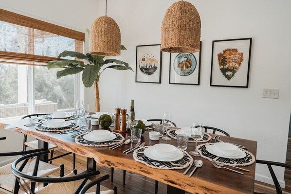 Un comedor con una mesa de madera dispuesta para seis personas, rodeada de sillas. Dos lámparas colgantes tejidas están encima de la mesa, y hay obras de arte enmarcadas en la pared.
