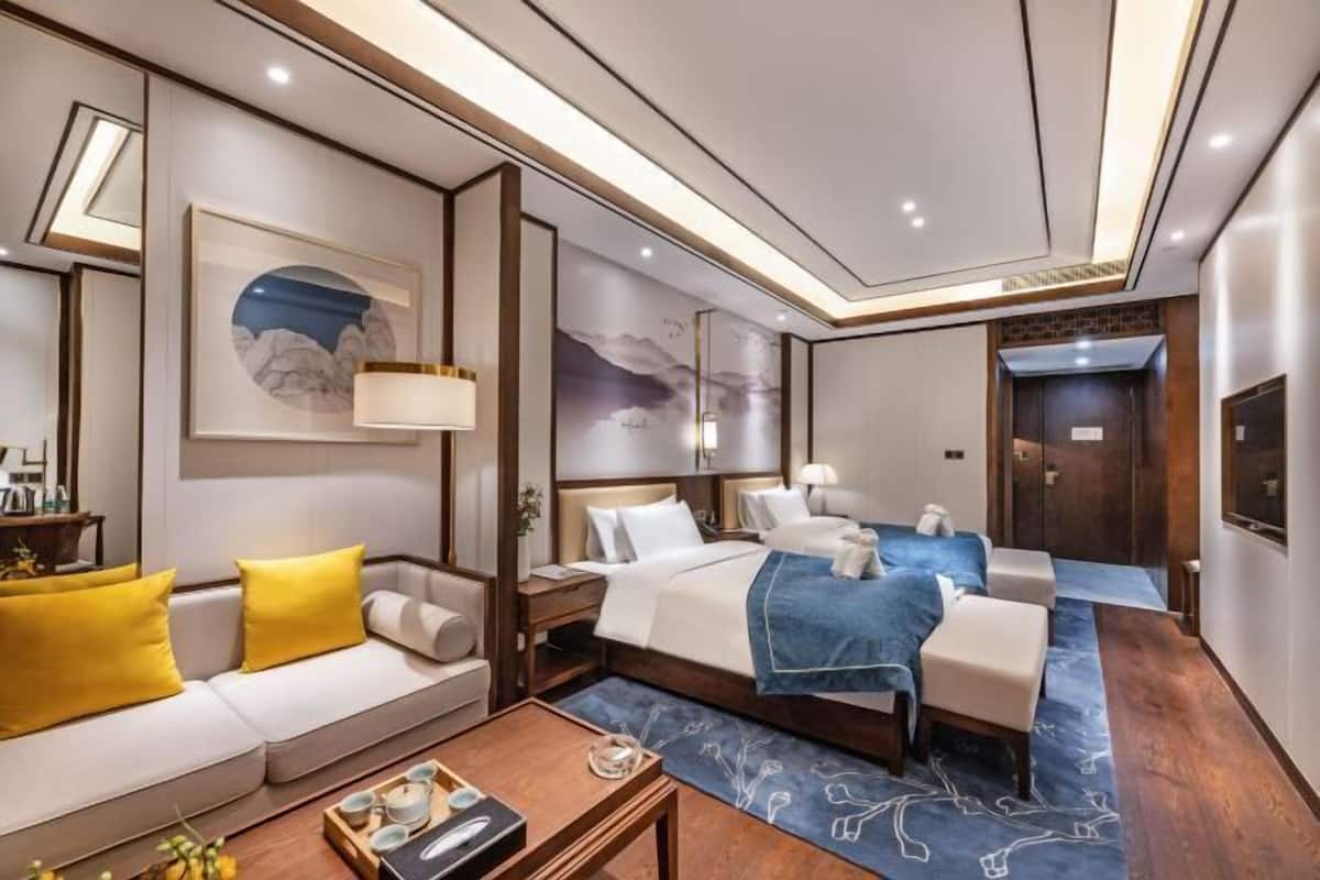 Habitación de hotel con cama, sofá con cojines amarillos, mesa de centro y obras de arte en las paredes.