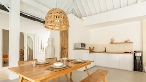 A spacious dining area with a wooden table set for four, a woven pendant light, and a kitchen in the background. - Villa Casa Bocami Canggu By Azure Near Finns (Canggu)