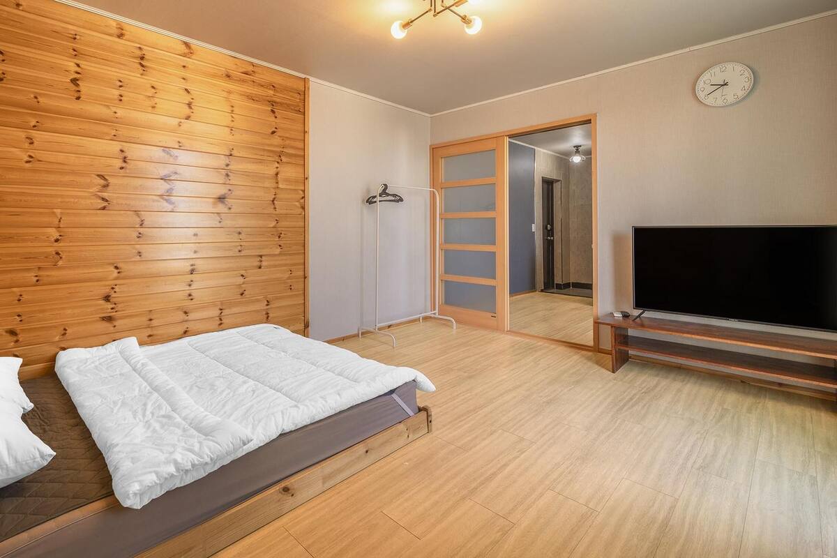 A bedroom with a wooden headboard, a bed with white bedding, a TV on a wooden stand, and a clock on the wall.