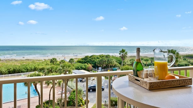 Een balkon met een ontbijtset, uitkijkend op een strand en een zwembad.